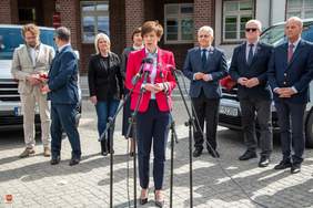 Pokaż zdjęcie: Konferencja prasowa przed budynkiem Starostwa Powiatowego w Pile. Na pierwszym planie kobieta o krótkich brązowych włosach przemawia do mikrofonów. Za nią stoi grupa siedmiu elegancko ubranych osób. Spotkanie dotyczy przekazania nowych busów dla Domów Pomocy Społecznej.