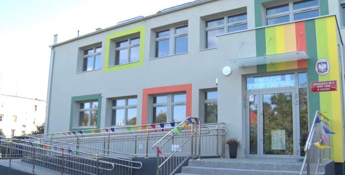 Pokaż zdjęcie: Dwukondygnacyjny nowoczesny budynek przedszkolny o jasnej elewacji z żywymi, kolorowymi obramowaniami wokół okien. Główne wejście z podjazdem dla wózków ozdobiono kolorowymi chorągiewkami, a obok drzwi wiszą czerwone tablice urzędowe z godłem Polski.