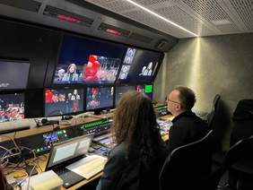 Pokaż zdjęcie: Zdjęcie konsoli i monitorów w studiu telewizyjnym. Przed monitorami siedzą operatorzy, kobieta i mężczyzna.
