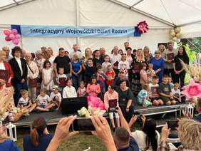 Pokaż zdjęcie: Uczestnicy Integracyjnego Wojewódzkiego Dnia Rodzin pozują do wspólnego zdjęcia na scenie pod namiotem. Wśród zgromadzonych są rodziny z dziećmi oraz osoby z pamiątkowymi dyplomami.