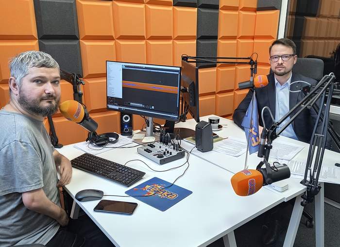 Pokaż zdjęcie: Dwóch mężczyzn siedzi w studiu radiowym przed mikrofonami z logo "Eska". Jeden z nich ma brodę i szarą koszulkę, drugi jest elegancko ubrany w garnitur i okulary. Przed nimi znajdują się komputery, sprzęt audio i notatki. Pomieszczenie jest wyłożone pomarańczowymi panelami akustycznymi, a zza szyby widać odbicie wnętrza studia.