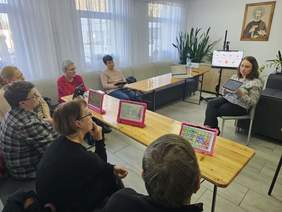 Kilka osób siedzi za biurkami, przed nimi siedzi kobieta, opowieada, prezentuje sprzęt