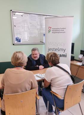 Pokaż zdjęcie: Mężczyzna siedzący przy stole rozmawia z kobietami, które są zwrócone tyłem do aparatu. Na ścianie tablica z licznymi informacjami. Za mężczyzną baner promujący PFRON