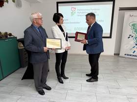Trzy osoby stoją przed ekranem z logo PFRON. Mężczyzna po lewej trzyma oprawiony dyplom, kobieta pośrodku ma biały żakiet, a mężczyzna po prawej trzyma grawerton w czerwonej ramie.  
