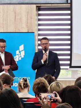 Przedstawiciel PFRON w granatowym garniturze przemawia do uczestników forum, trzymając mikrofon. W tle widoczny jest baner wydarzenia oraz drugi prelegent.