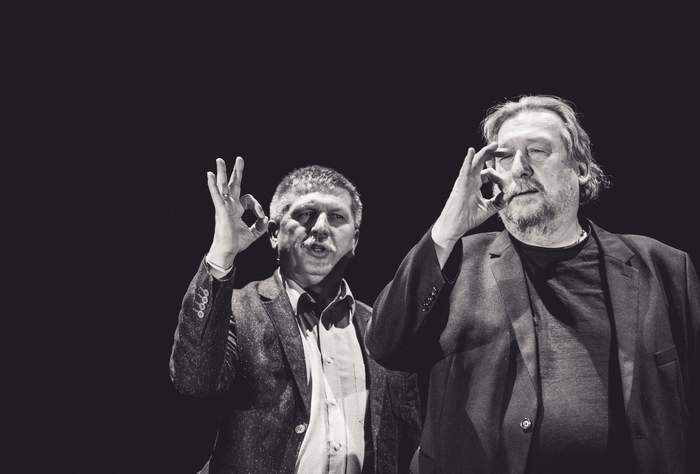 Pokaż zdjęcie: na czarnym tle dwaj mężczyźni stoją obok siebie wykonując ruchy rąk uczą się języka migowego