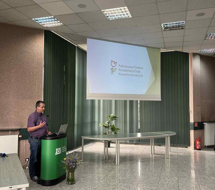 Pokaż zdjęcie: Przestronna sala konferencyjna z ekranem emitującym logo PFRON. Z lewej strony przemawiający do mikrofonu prelegent. Stoi za mównicą z logo ZUS.