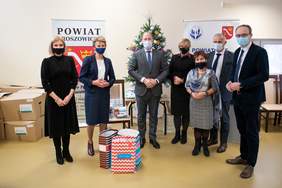 Pokaż zdjęcie: Pierwszy plan - siedem osób. Kobiety i mężczyźni elegancko ubrani, w maseczkach. W tle pudełka oznakowane logotypem PFRON, przygotowane do przekazania osobom potrzebującym.