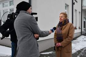 Pokaż zdjęcie: Reporter przeprowadza wywiad z kobietą blondynką w brązowym płaszczu. Stoją na parkingu Centrum Opiekuńczo Mieszkalnego w Jaszczowie.