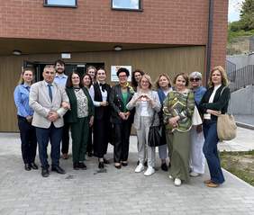 Pokaż zdjęcie: Grupowe zdjęcie kilkunastu osób przed nowoczesnym budynkiem z cegły i drewna. Wszyscy uśmiechają się, kobieta w środku pokazuje znak serca. Nad wejściem widoczny szyld placówki.