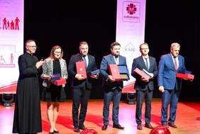 Pokaż zdjęcie: a scenie znajduje się sześć osób, w tym ksiądz w sutannie oraz pięć osób ubranych elegancko, trzymających nagrody. W tle widoczne są logotypy Caritas oraz innych organizacji związanych z wydarzeniem.