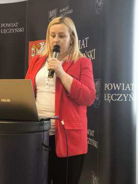 Pokaż zdjęcie: Kobieta w czerwonej marynarce trzyma w ręku mikrofon.
