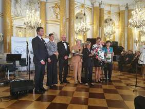 Pokaż zdjęcie: Grupa ludzi elegencko ubranych, w pięknym stylowym, zabytkowym miejscu, złocienia na ścianach, pozuje do zdjęcia ze statuetkami Lodołamaczy. 