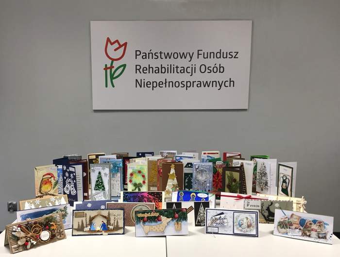 Pokaż zdjęcie: Na zdjęciu w środkowej jego części widać logo Państwowego Funduszu Rehabilitacji Osób Niepełnosprawnych. Poniżej na jasnym stole ustawione jest kilkadziesiąt kartek świątecznych ułożonych w kilku rzędach.  Kartki są różnego koloru i przedstawiają motywy choinek, gwiazd świątecznych dekoracji i zimowych ilustracji. Kartki świąteczne zostały nadesłane na Konkurs kartek bożonarodzeniowych.