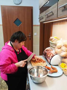 Pokaż zdjęcie: W kuchni znajduje się kobieta. Kroi na desce kiełbaski. Ma założony czarny fartuch. Na blacie kuchennym leży pieczywo. 