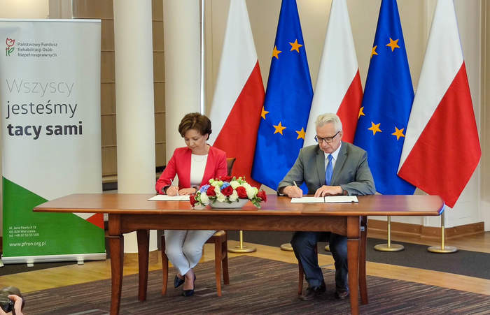 Pokaż zdjęcie: Kobieta i mężczyzna siedzą przy biurku i podpisują dokumenty. Na stole stoi bukiet kwiatów, w tle flagi polskie i unii europejskiej
