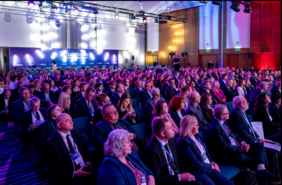 Pokaż zdjęcie: Duża konferencja w sali z oświetleniem scenicznym. Publiczność siedzi w rzędach, uczestnicy w eleganckich strojach słuchają wystąpienia. W tle scena z ekranem i kamerami rejestrującymi wydarzenie.