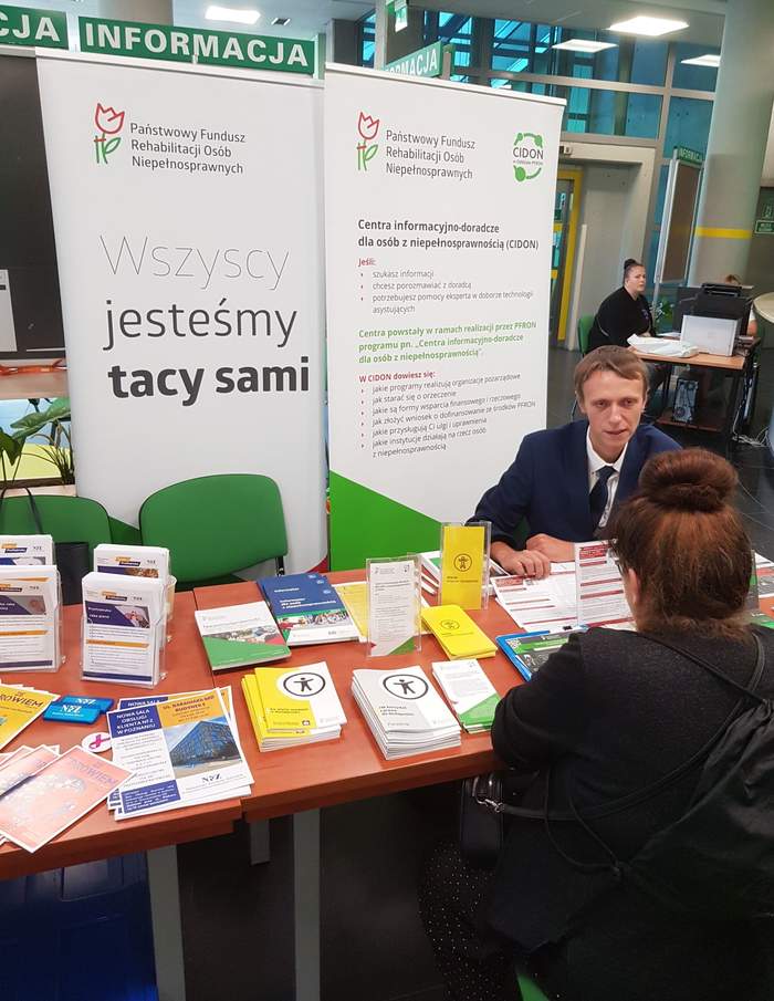 Pokaż zdjęcie: Mężczyzna siedzi za stolikiem na tle baneru PFRON oraz CIDON i udziela informacji kobiecie, która przyszła do ZUS na Dzień Osób z Niepełnosprawnością. Na stoliku ułożone są materiały informacyjne promujące programy i zadania PFRON.
