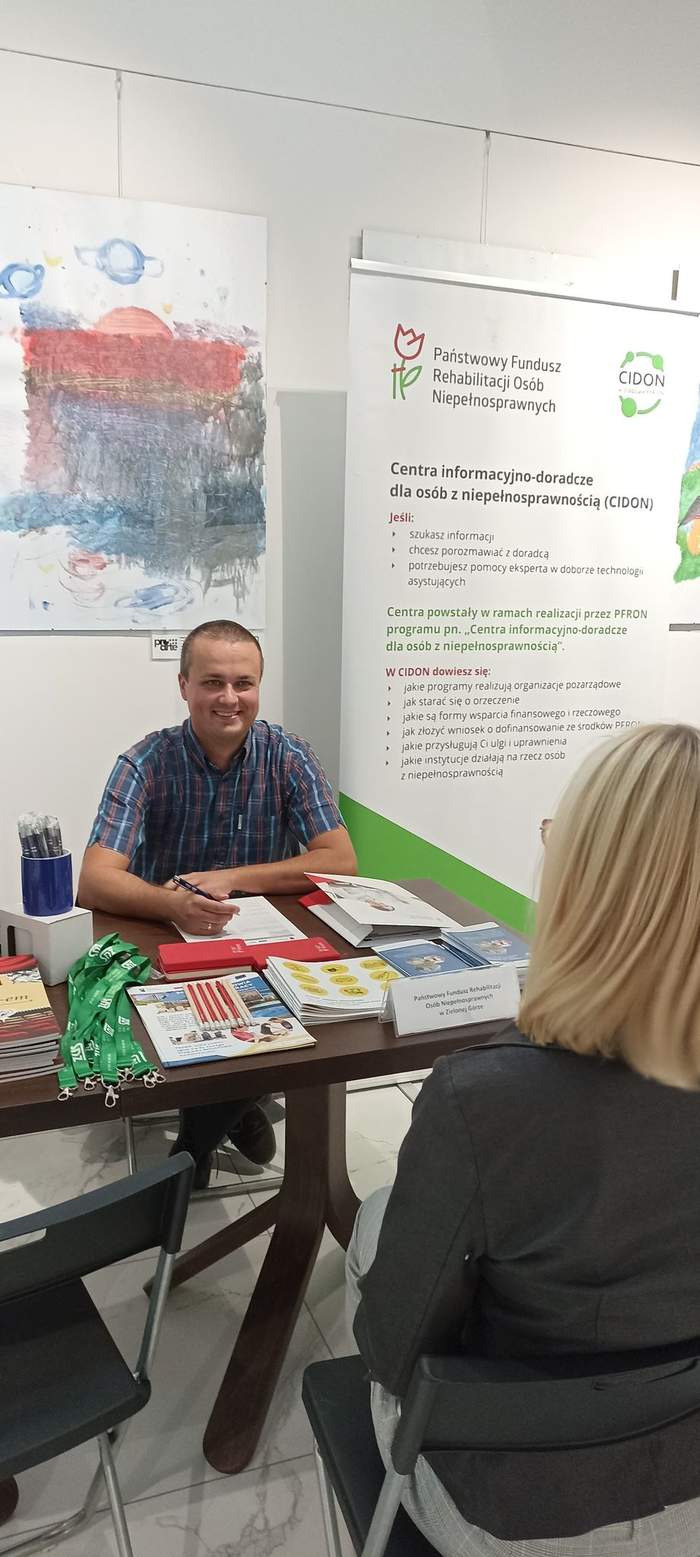 Pokaż zdjęcie: Pracownik Oddziału PFRON w Zielonej Górze siedzi przy stoliku na tle roll-up PFRON i rozmawia z klientką