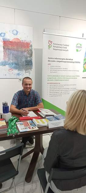 Pokaż zdjęcie: Pracownik Oddziału PFRON w Zielonej Górze siedzi przy stoliku na tle roll-up PFRON i rozmawia z klientką