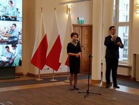 Pokaż zdjęcie: przemawiająca kobieta, w tle biało-czerwone flagi, obok mężczyzna - tłumacz języka migowego