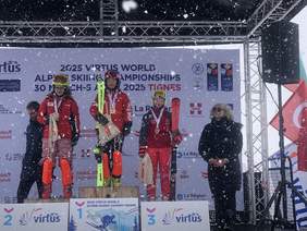 Trzech narciarzy na podium medalowym, dodatkowo widoczne dwie osoby kobieta po prawej i z tyłu mężczyzna. Zimowa aura, sypiący śnieg.