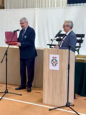 Pokaż zdjęcie: Dwóch mężczyzn stoi w szkolnej sali gimnastycznej. Przed nimi stoją mikrofony. Jeden z panów prezentuje pamiątkowy grawer z dedykacją. 