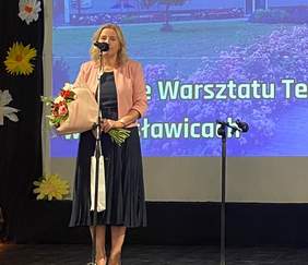 Pokaż zdjęcie: Na scenie przed mikrofonem stoi kobieta ubrana w granatową sukienkę i łososiową marynarkę. W ręku trzyma bukiet kwiatów.  