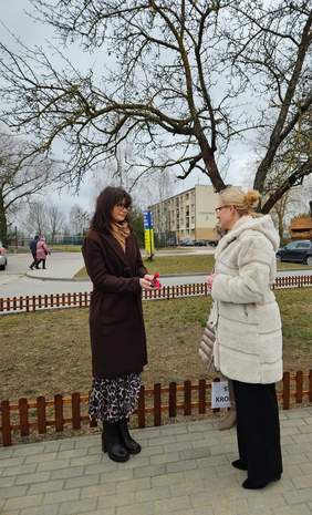 Pokaż zdjęcie: Na zdjęciu dwie kobiety stoją na chodniku, jedna z nich wręcza drugiej kluczyki. Kobieta po lewej ubrana jest w brązowy płaszcz i czarne buty, natomiast kobieta po prawej nosi jasny futrzany płaszcz i czarne spodnie. W tle widoczny jest budynek mieszkalny, kilka samochodów oraz drzewa bez liści. 