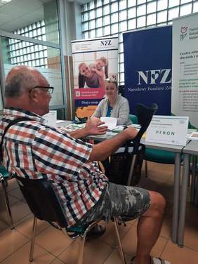 Pokaż zdjęcie: W pomieszczeniu Oddziału ZUS ustawione są stoły. Przy jednym z nich naprzeciwko siebie siedzą kobieta i mężczyzna. Rozmawiają ze sobą. W tle widać baner PFRON oraz NFZ. 