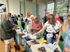 Pokaż zdjęcie: Przy stolikach na których leża materiały promocyjne i informatory stoją przedstawiciele instytucji, naprzeciw stoją osoby które sięgają po materiały promocyjne.