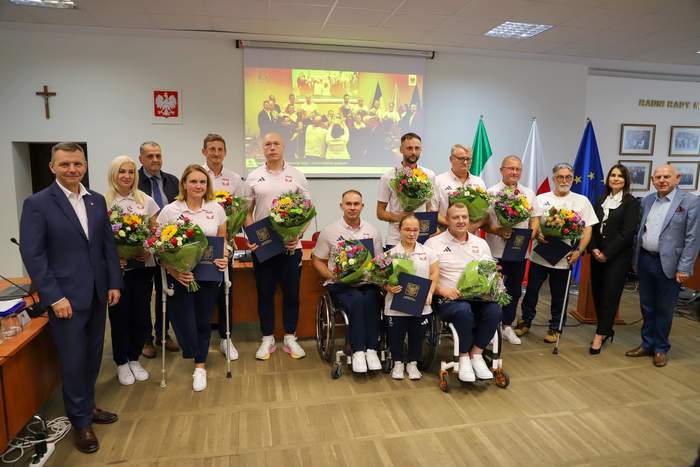 Pokaż zdjęcie: Paralimpijczycy na wspólnym zdjęciu, trzymają w rękach bukiety, są ubrani na sportowo, są przedstawiciele władz miasta