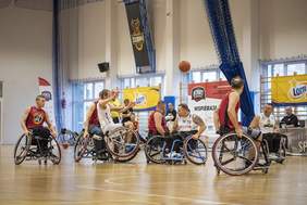 Pokaż zdjęcie: Zawodnicy koszykówki na wózkach podczas meczu na hali sportowej, widoczni w dynamicznej akcji. W tle banery sponsorów oraz logo organizatora – START Białystok.