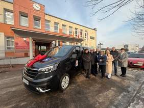 Pokaż zdjęcie: Na pierwszym planie po lewej stronie stoi czarny van z dużą czerwoną kokardą na masce. Na przednich drzwiach pojazdu widoczny jest herb Gminy Czerniejewo. Przed maską na ziemi znajdują się mokre i częściowo oblodzone nawierzchnie. Po prawej stronie samochodu stoi grupa dziewięciu osób uśmiechniętych, ubrane w ciemne i jasne płaszcze zimowe. Za nimi, w tle, widać dwukondygnacyjny budynek w kolorze żółto-pomarańczowym z zegarem na elewacji nad wejściem i flagami przy wejściu.