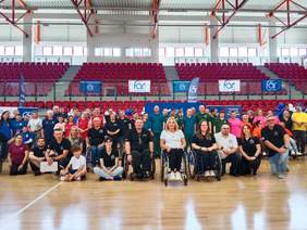Pokaż zdjęcie: W hali sportowej stoi tłum ludzi. Wszyscy są ustawieni w rzędach i pozują do zdjęcia. W tle widać trybuny. 