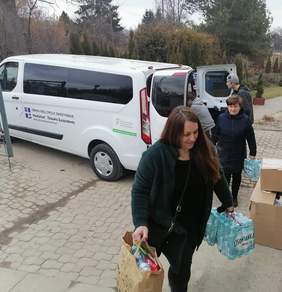 dwie kobiety niosą w rękach m.in. wodę, za nimi widać białego busa obok którego stoi mężczyzna
