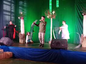 Pokaż zdjęcie: Na scenie stoi chłopak, który śpiewa do mikrofonu. W tle tańczą inne osoby. Są także dekoracje sceniczne - sztuczne drzewa. 