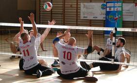 Pokaż zdjęcie: Siędzący na boisku zawodnicy w strojach sportowych przedzieleni nisko zawieszoną siatką, w powietrzu znajduje się rzucona piłka do siatkówki