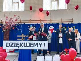 Pokaż zdjęcie: W szkolnej sali gimnastycznej ustawiona jest scena. Stoi na niej dyrektor Łódzkiego Oddziału PFRON, który odbiera nagrodę "Kryształowe Serce". 