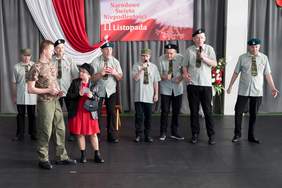 Pokaż zdjęcie: Grupa osób w wojskowych stylizacjach stoi na scenie podczas uroczystości 11 listopada; część z nich trzyma mikrofony, a w tle widoczne są biało-czerwone dekoracje i plakat z napisem „Narodowe Święto Niepodległości 11 Listopada”.