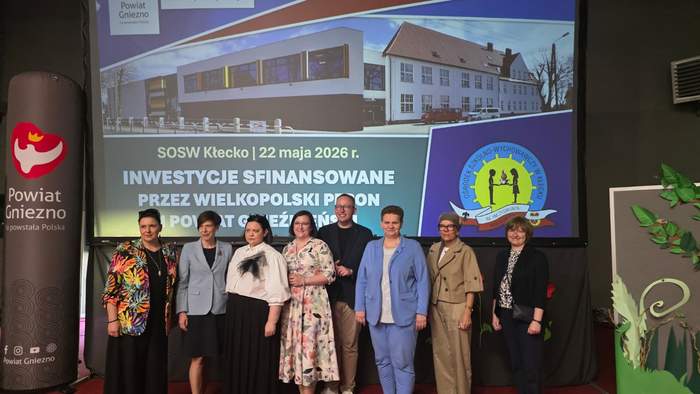 Pokaż zdjęcie: Grupa ośmiu osób stoi w jednym rzędzie na czerwonej scenie na tle dużego ekranu multimedialnego. Na slajdzie widoczna jest wizualizacja budynku oraz napisy: „SOSW Kłecko i  i data 22 maja 2026 roku, a poniżej napis: „Inwestycje sfinansowane przez wielkopolski PFRON”. Po lewej stronie sceny stoi pionowy baner Powiatu Gniezno z czerwonym herbem, a po prawej dekoracyjny panel z motywem zielonych liści.