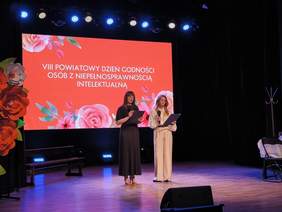 Pokaż zdjęcie: Dwie osoby stoją na scenie i czytają z kart, trzymając mikrofony. Za nimi duży ekran z napisem „VIII Powiatowy Dzień Godności Osób z Niepełnosprawnością Intelektualną” oraz dekoracją kwiatową i oświetleniem sceny.