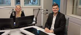 Pokaż zdjęcie: Studio radiowe. Przy białym stole z platformami siedzi pan dyrektor. Dziennikarka siedzi przy konsoli z dwoma monitorami. Z jej biurka na długich metalowych stelażach wychodzą dwa mikrofony skierowane w stronę rozmówców