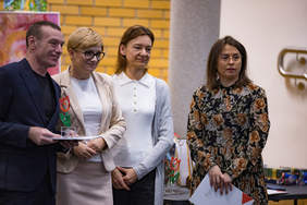 Pokaż zdjęcie: Cztery osoby stoją obok siebie. Mężczyzna po lewej stronie trzyma dyplom i statuetkę w kształcie tulipana. Obok niego stoją trzy kobiety – jedna z dokumentami w rękach. Wszyscy pozują do zdjęcia podczas gali konkursowej.