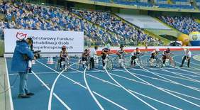 Pokaż zdjęcie: Na stadionie przy pełnych trybunach siedmiu sportowców szykuje się do startu w wyścigu rowerowym. W tle logo PFRON. Wydarzenie jest transmitowane przez stojącego z lewej strony kamerzystę. Z prawej strony stoi sędzia startowy.