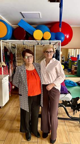 Pokaż zdjęcie: Po prawej stronie stoi Dyrektor Oddziału Podlaskiego PFRON Edyta Kondzior, po lewej Irena Świacka – Sekretarz Zarządu Koła Polskie Stowarzyszenie na rzecz Osób z Niepełnosprawnością Intelektualną w Suwałkach; kobiety znajdują się w sali rehabilitacyjnej, w tle widoczny sprzęt do ćwiczeń.