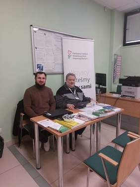 Pokaż zdjęcie: Dwóch mężczyzn siedzi przy stole, na którym znajdują się informatory, broszury, ulotki i poradniki. Za nimi tablica z ogłoszeniami oraz roll-up promujący PFRON. Po drugiej stronie stołu dwa krzesła. Z prawej strony fotografii biurko z komputerem oraz drukarką. Znajdują się w pomieszczeniu.