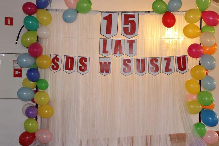 Pokaż zdjęcie: NA zdjęciu czerwony baner z napisem 15 lat ŚDS w Suszu, wokół niego kolorowe balony
