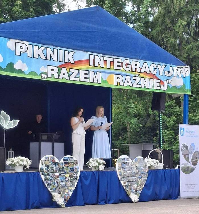 Pokaż zdjęcie: Scena plenerowa pod niebieskim namiotem; dwie osoby czytają z kartki, nad nimi baner „Piknik Integracyjny Razem Raźniej”, przed sceną dekoracje i kolaże zdjęć.