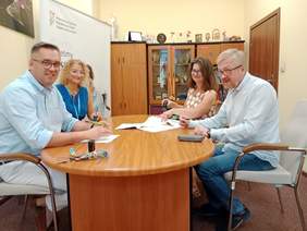 Pokaż zdjęcie: Przy owalnym stole siedzą cztery osoby: dwie kobiety i dwóch mężczyzn. Podpisują umowę. Na stole leżą różne dokumenty, pieczątki, okulary i klucze. Wszyscy się uśmiechają. 